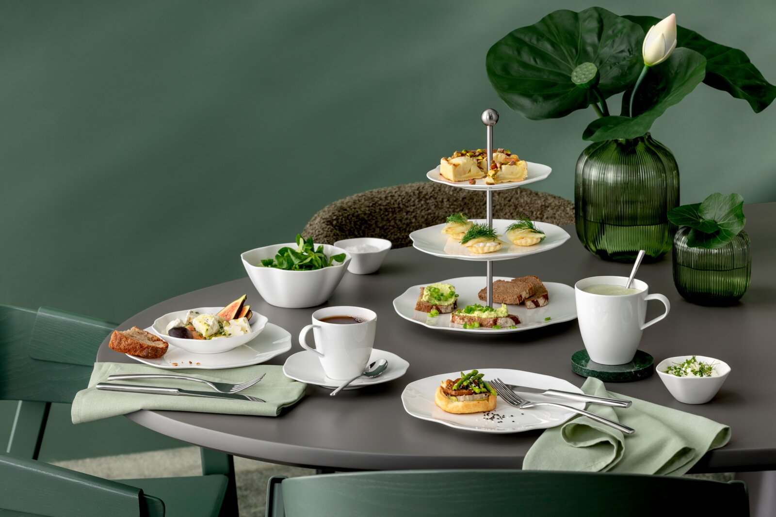 Schräg von vorne fotografierter gedeckter Tisch mit weißem Porzellan und zweistöckiger Etagere mit kleinen Häppchen, Tassen und Schalen sowie grünen Glasvasen im Hintergrund