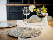 Modern gedeckter Esstisch mit großem Teller im Mosaik-Design, zwei Weingläsern (eins mit Rotwein) und weißer Vase mit Blumen, fotografiert aus leicht erhöhter, schräger Frontperspektive.