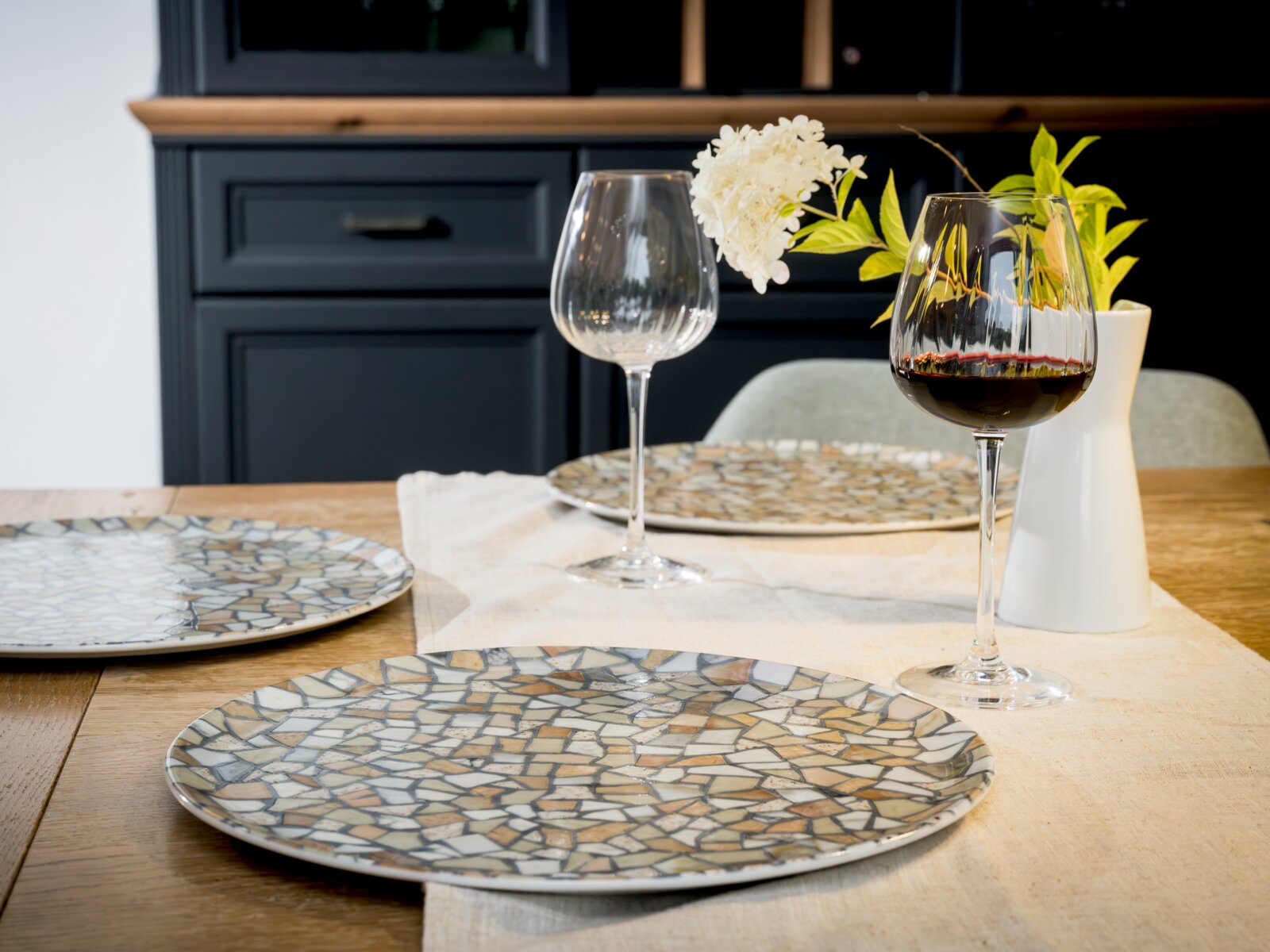 Modern gedeckter Esstisch mit großem Teller im Mosaik-Design, zwei Weingläsern (eins mit Rotwein) und weißer Vase mit Blumen, fotografiert aus leicht erhöhter, schräger Frontperspektive.