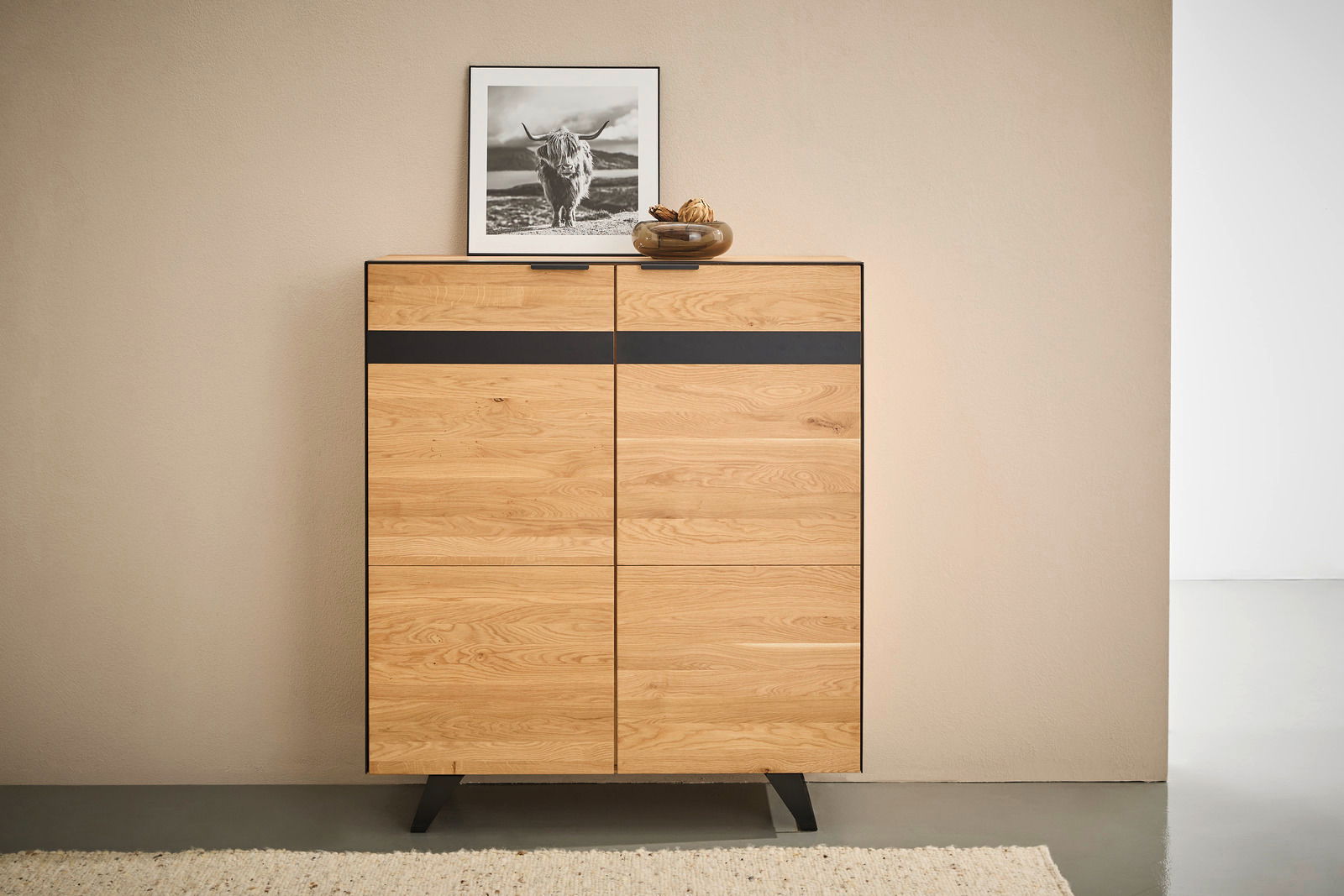 Frontale Ansicht eines modernen Highboards aus Holz mit schwarzem Akzentstreifen, dekoriert mit einem Bild und einer Schale.