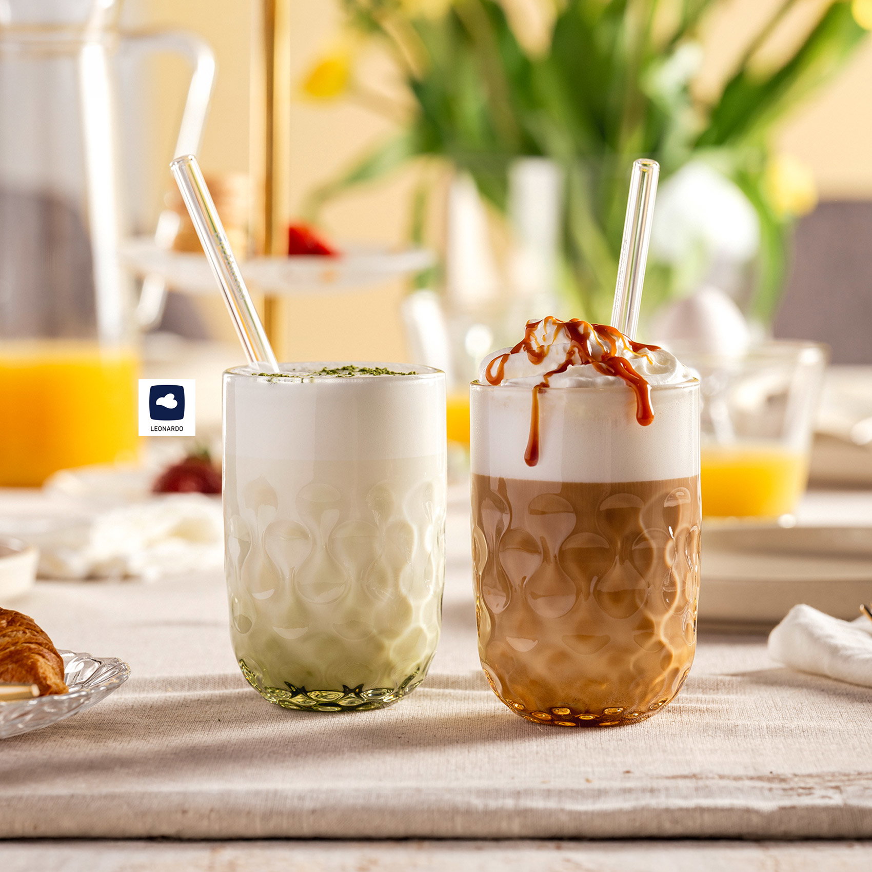 Zwei Latte-Getränke im strukturierten Glas – Matcha Latte und Caramel Latte mit Sahne – auf gedecktem Frühstückstisch mit Croissant und Orangensaft
