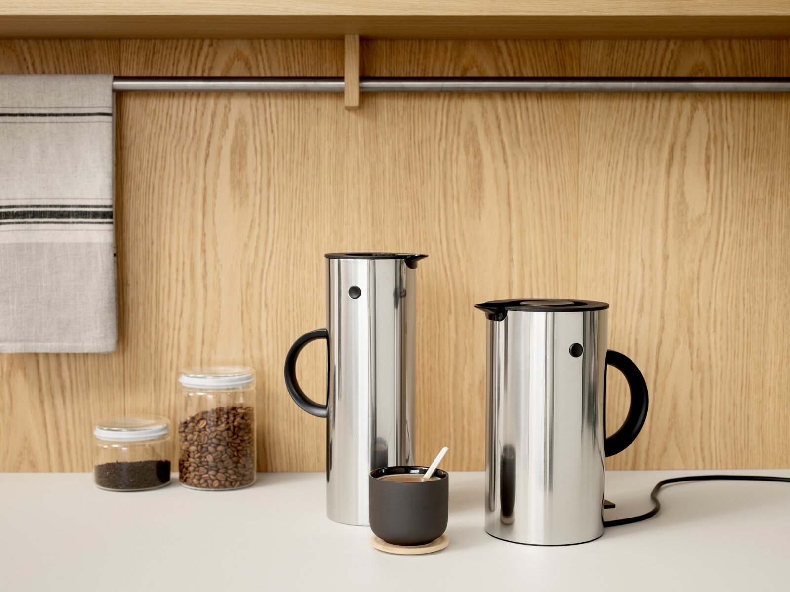 Stelton Thermoskan Twee elegante thermoskannen van staal met een zwarte handgreep, op een aanrechtblad voor een houten wand, gefotografeerd vanuit een zijaanzicht.