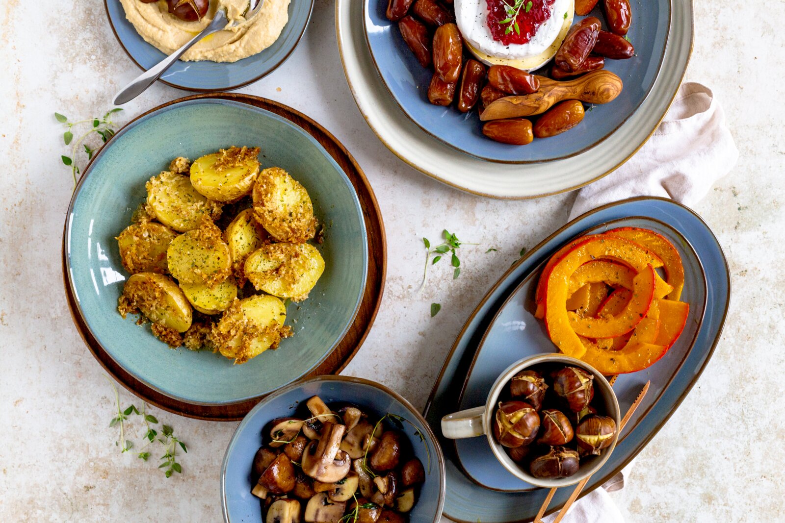 Draufsicht auf ein herbstliches Food-Arrangement: gewürzte Kartoffeln in einer Schüssel, gebratene Pilze, geschnittenen Kürbis und geröstete Kastanien sowie Datteln mit cremigem Dip auf blauem Geschirr.