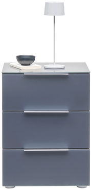 Moderne Nachtkonsole mit drei Schubladen in grauer Farbe, darauf eine weiße Tischlampe, eine Schale und Bücher. Frontalansicht.