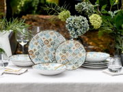 Bunt gemustertes Keramik-Geschirrset mit zwei Speisetellern und mehreren Schalen auf weiß gedecktem Tisch mit Gläsern und Blumen, frontal in leichter Schrägansicht aufgenommen