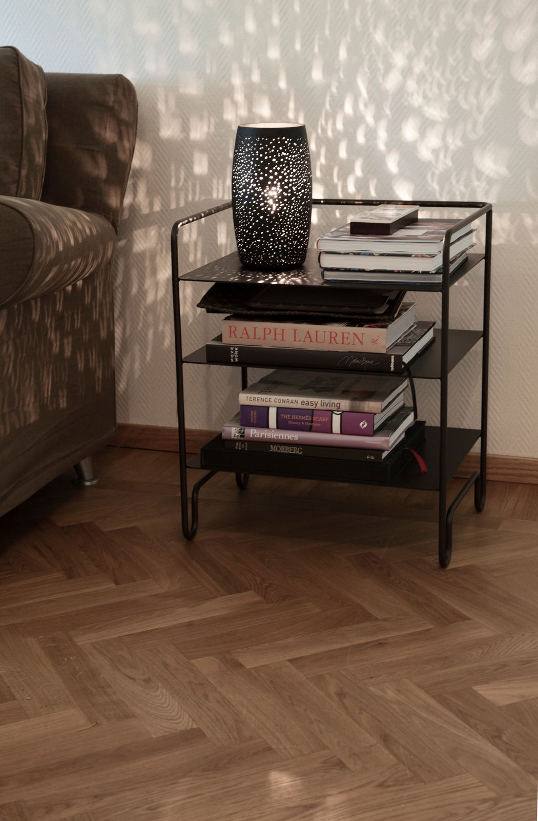 Tischleuchte Colby in sand-schwarz auf einem Regal, seitliche Perspektive, mit dekorativem Lichtmuster an der Wand.