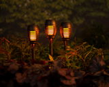 Drei solarbetriebene Fackeln aus Plastik mit flackerndem Licht, in einem Garten bei Dämmerung, aus der Perspektive von vorne aufgenommen.
