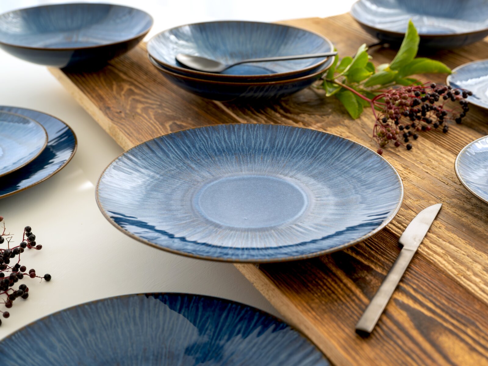 Blau glasierter Keramik-Essteller mit radialem Muster auf rustikalem Holztisch, umgeben von passenden Schalen und Besteck, schräg von oben fotografiert.