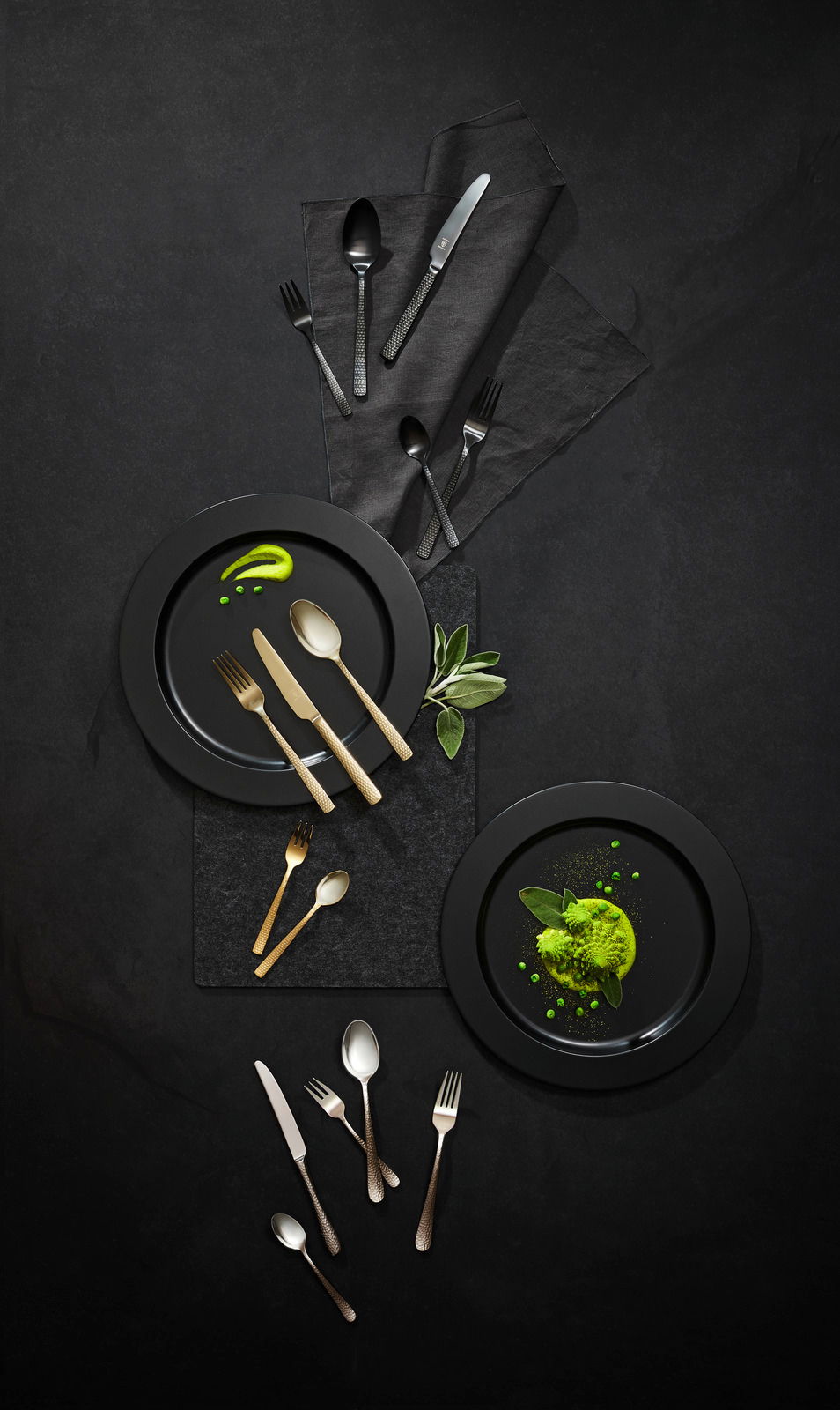 Vogelperspektive auf ein stilvolles Besteckset 'Sunset' in Gold und Schwarz, arrangiert auf dunklen Tellern mit grüner Dekoration.