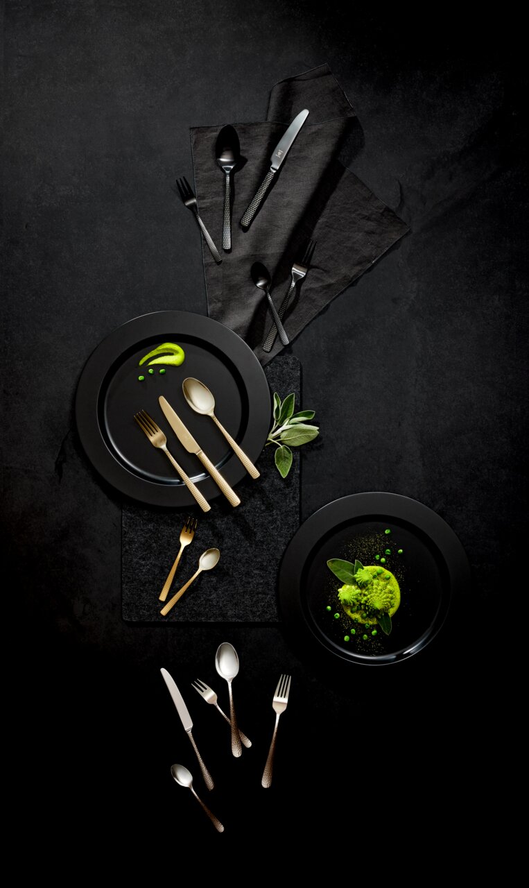 Vogelperspektive auf ein stilvolles Besteckset 'Sunset' in Gold und Schwarz, arrangiert auf dunklen Tellern mit grüner Dekoration.