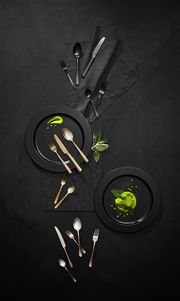 Vogelperspektive auf ein stilvolles Besteckset 'Sunset' in Gold und Schwarz, arrangiert auf dunklen Tellern mit grüner Dekoration.