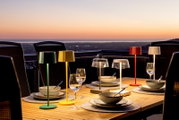 Mehrfarbige Akku-Tischleuchten auf einem gedeckten Tisch bei Sonnenuntergang, seitliche Perspektive.