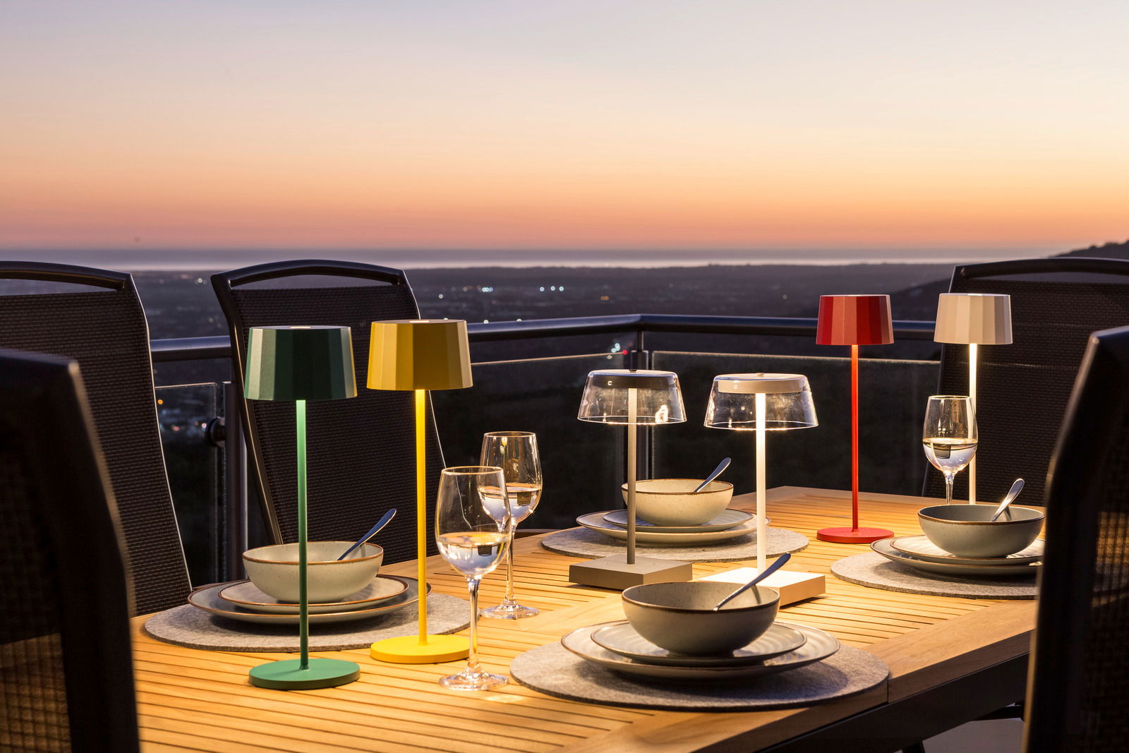 Mehrfarbige Akku-Tischleuchten auf einem gedeckten Tisch bei Sonnenuntergang, seitliche Perspektive.