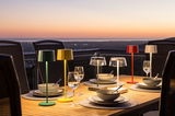 Mehrfarbige Akku-Tischleuchten auf einem gedeckten Tisch bei Sonnenuntergang, seitliche Perspektive.