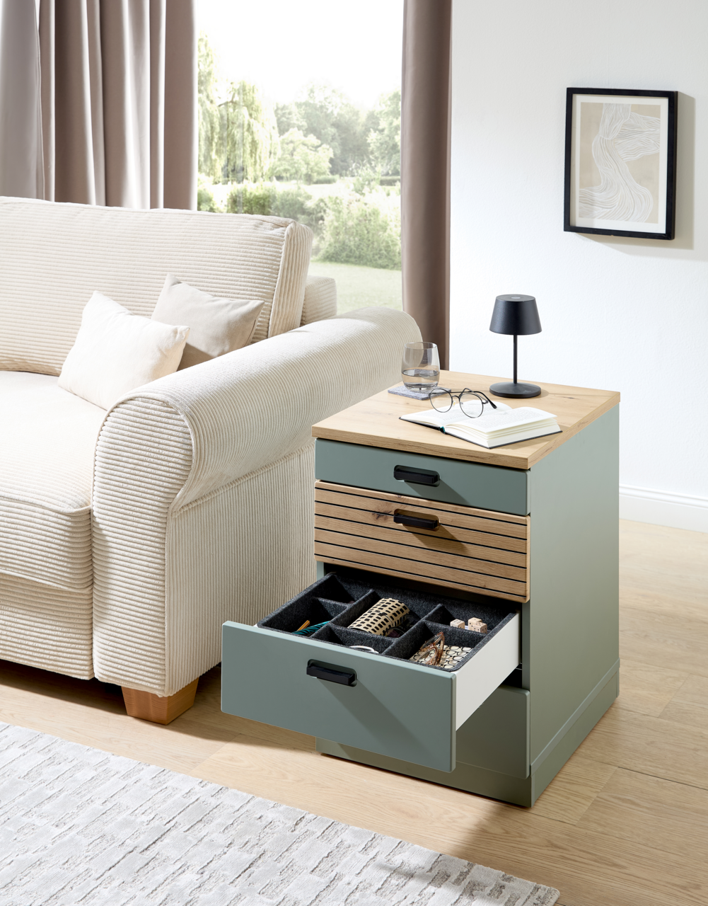 Rollcontainer mit drei Schubladen, teilweise geöffnet, in einem modernen Wohnzimmer neben einem Sofa, aus seitlicher Perspektive fotografiert.