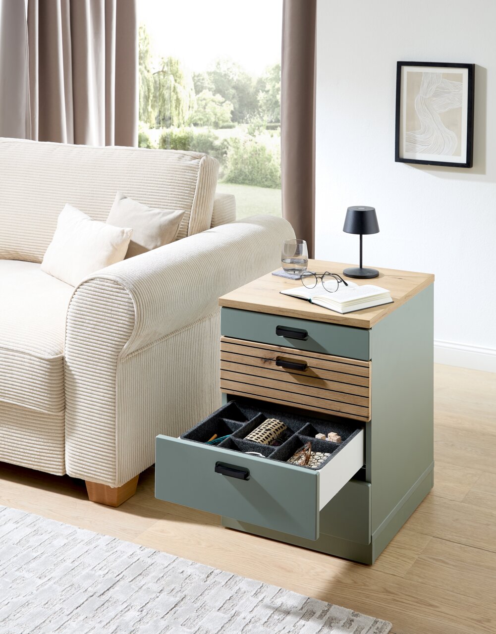 Rollcontainer mit drei Schubladen, teilweise geöffnet, in einem modernen Wohnzimmer neben einem Sofa, aus seitlicher Perspektive fotografiert.