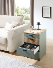 Rollcontainer mit drei Schubladen, teilweise geöffnet, in einem modernen Wohnzimmer neben einem Sofa, aus seitlicher Perspektive fotografiert.