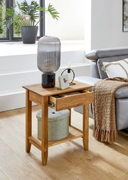 Moderner Beistelltisch aus hellem Holz mit Schublade und Ablagefach, darauf geriffelte Glas-Tischlampe und kleiner Wecker, im Wohnzimmer neben grauem Sofa; Perspektive: leicht erhöhte Frontansicht schräg von links.