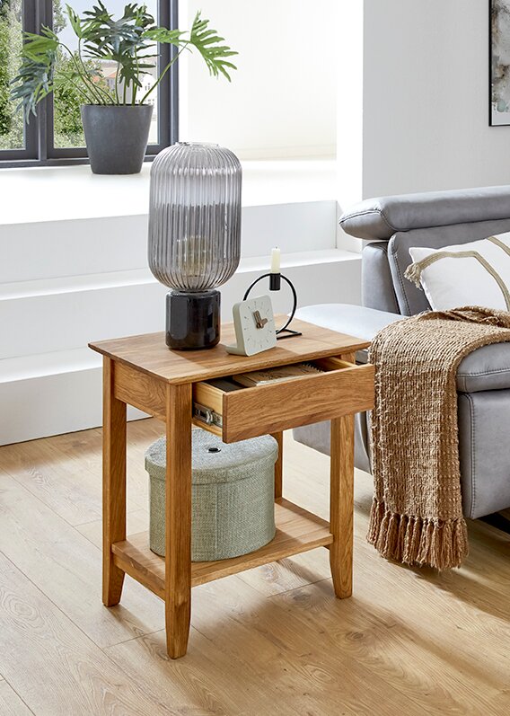 Moderner Beistelltisch aus hellem Holz mit Schublade und Ablagefach, darauf geriffelte Glas-Tischlampe und kleiner Wecker, im Wohnzimmer neben grauem Sofa Perspektive: leicht erhöhte Frontansicht schräg von links.