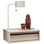 Beiger Nachttisch mit Schublade und schwarzem Griff, darauf eine weiße Tischlampe sowie braune Glasflasche und Glas auf Tablett, frontal aus leicht erhöhter Perspektive