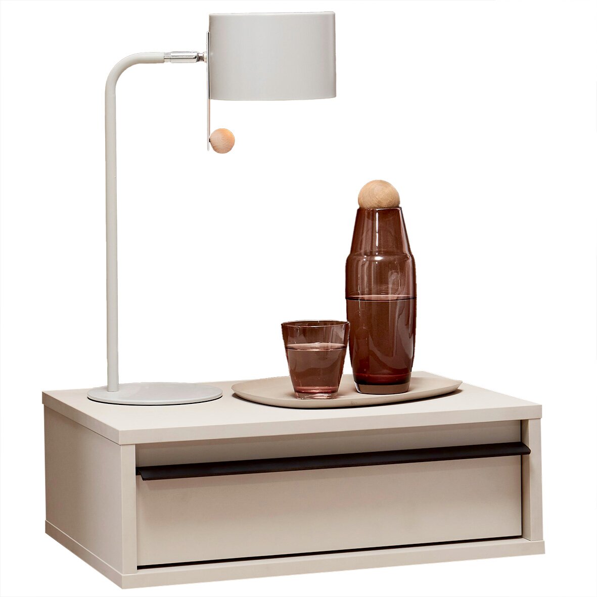 Beiger Nachttisch mit Schublade und schwarzem Griff, darauf eine weiße Tischlampe sowie braune Glasflasche und Glas auf Tablett, frontal aus leicht erhöhter Perspektive