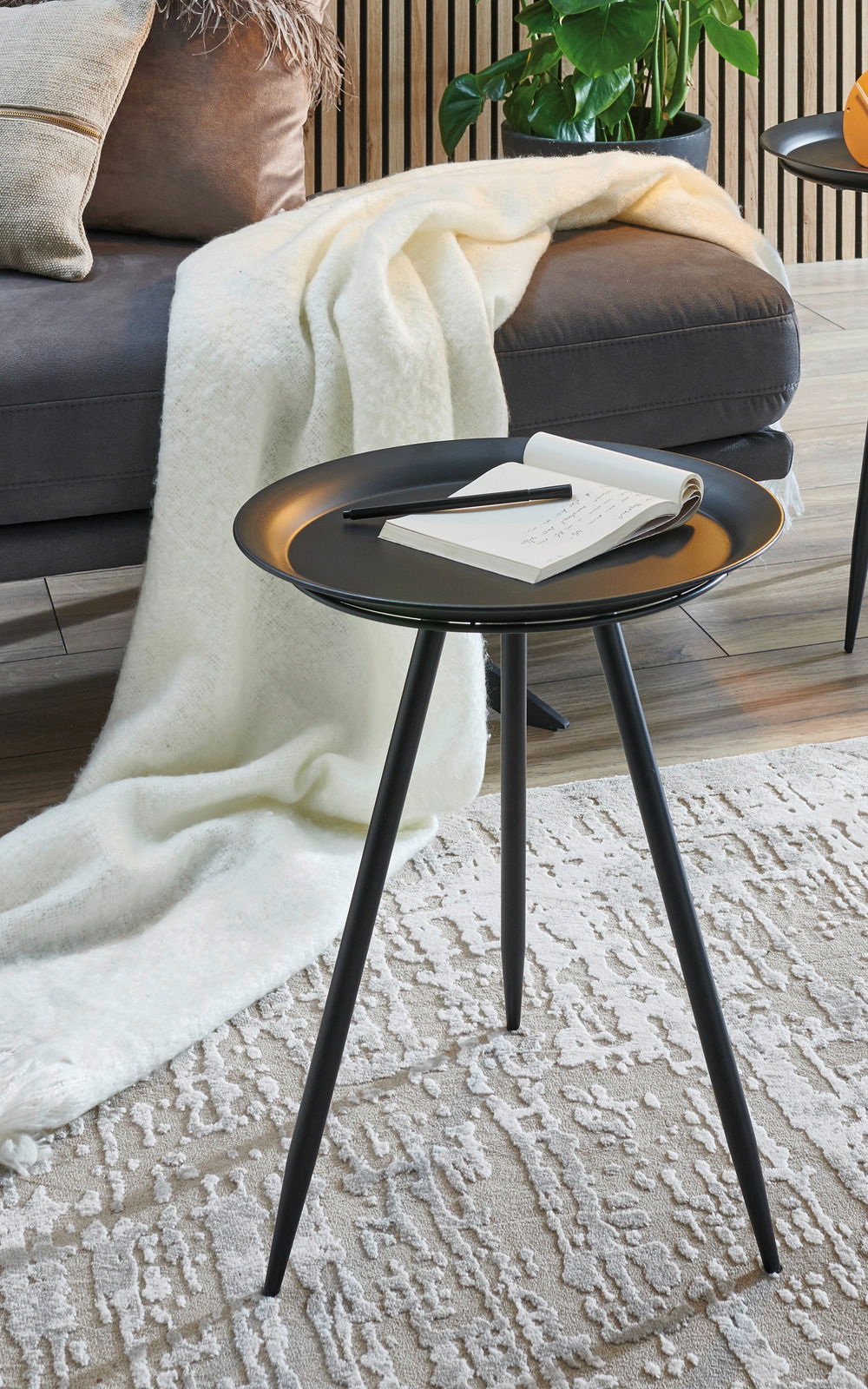 Moderner schwarzer Beistelltisch mit drei Beinen, von oben fotografiert, auf einem gemusterten Teppich neben einem Sofa mit Decke.