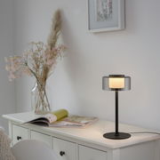 Moderne Tischleuchte RIKA mit schwarzem Standfuß und transparentem Lampenschirm, seitliche Perspektive auf einem weißen Schrank platziert.