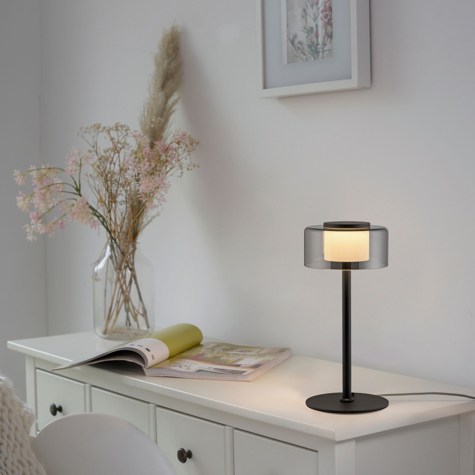 Moderne Tischleuchte RIKA mit schwarzem Standfuß und transparentem Lampenschirm, seitliche Perspektive auf einem weißen Schrank platziert.