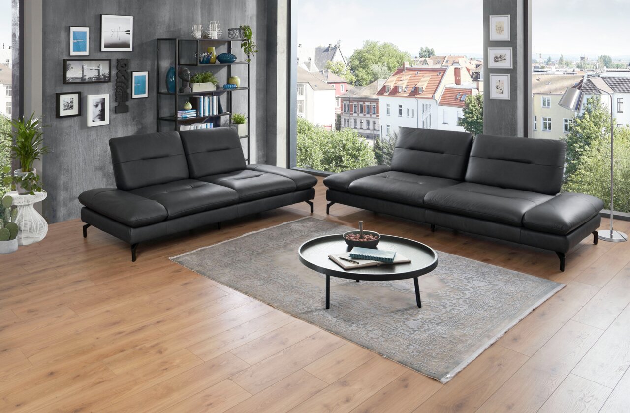 Moderano Sofa 2er ORLANDO Zwei moderne schwarze 2er-Sofas in einem stilvoll eingerichteten Wohnzimmer mit Holzboden und großen Fenstern, die einen Blick auf die Stadt bieten. Perspektive: Frontalansicht.