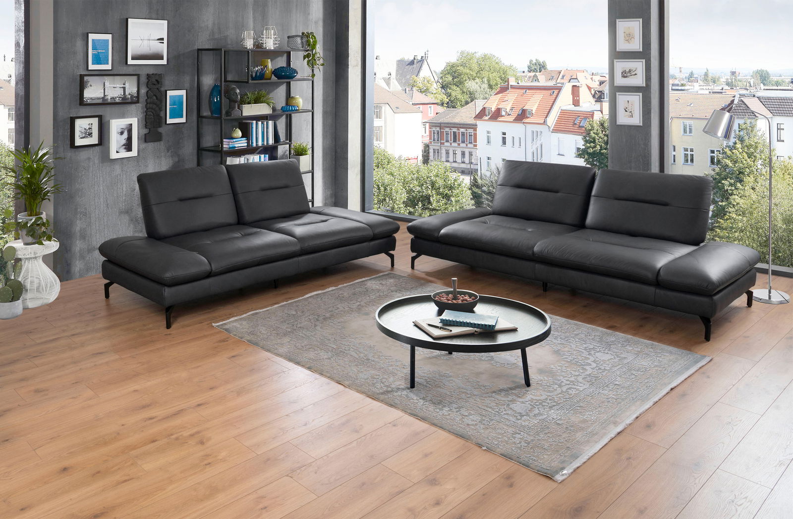 Zwei moderne schwarze 2er-Sofas in einem stilvoll eingerichteten Wohnzimmer mit Holzboden und großen Fenstern, die einen Blick auf die Stadt bieten. Perspektive: Frontalansicht.