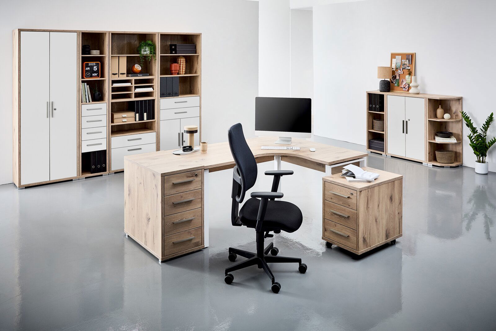 Moderne Büromöbel in einem hellen Raum, bestehend aus einem Eckschreibtisch mit Rollcontainer und einem schwarzen Bürostuhl. Im Hintergrund befinden sich Regale und Schränke mit Aktenordnern und Dekorationen. Perspektive: Frontalansicht.
