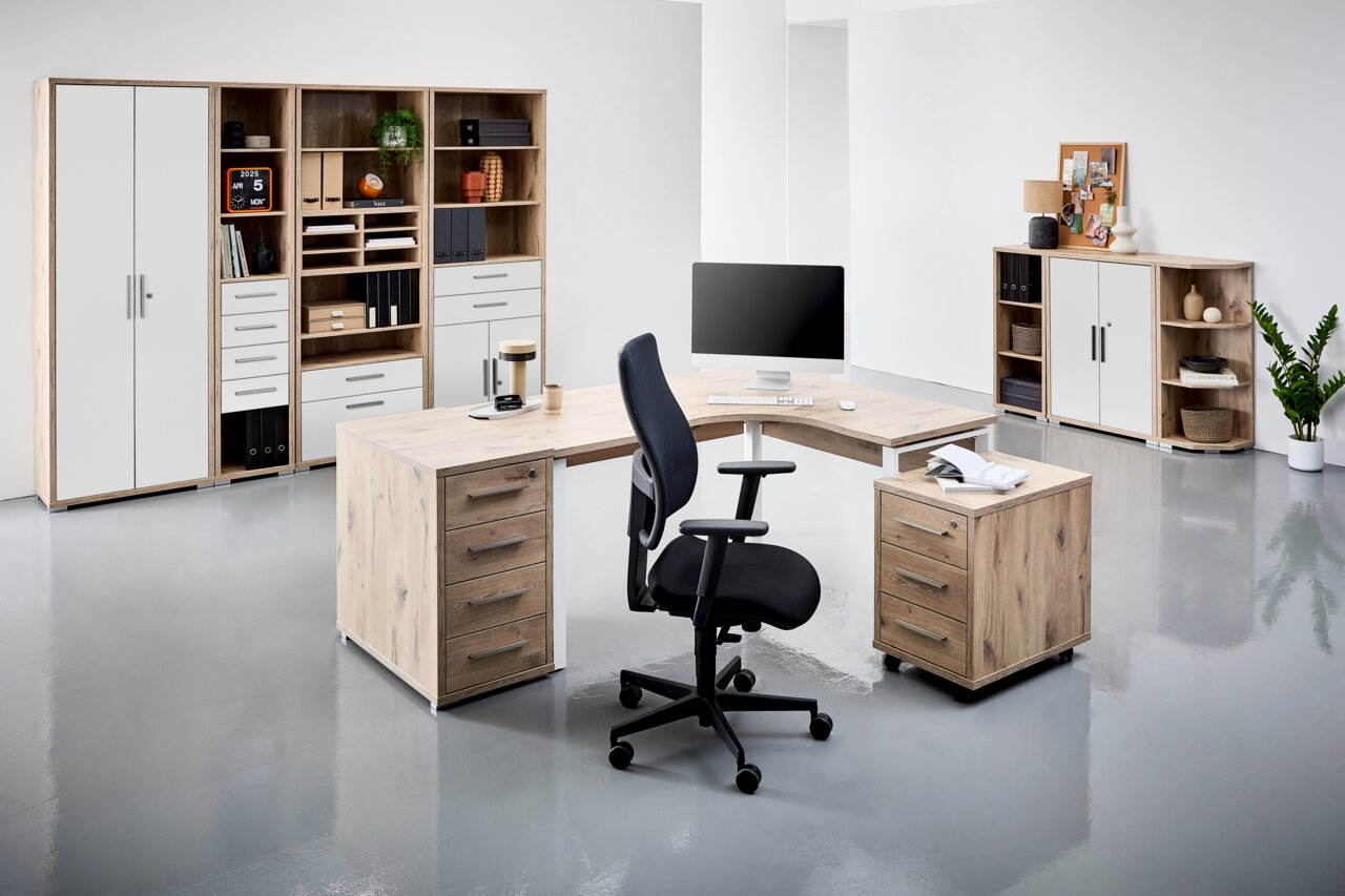 Moderne Büromöbel in einem hellen Raum, bestehend aus einem Eckschreibtisch mit Rollcontainer und einem schwarzen Bürostuhl. Im Hintergrund befinden sich Regale und Schränke mit Aktenordnern und Dekorationen. Perspektive: Frontalansicht.