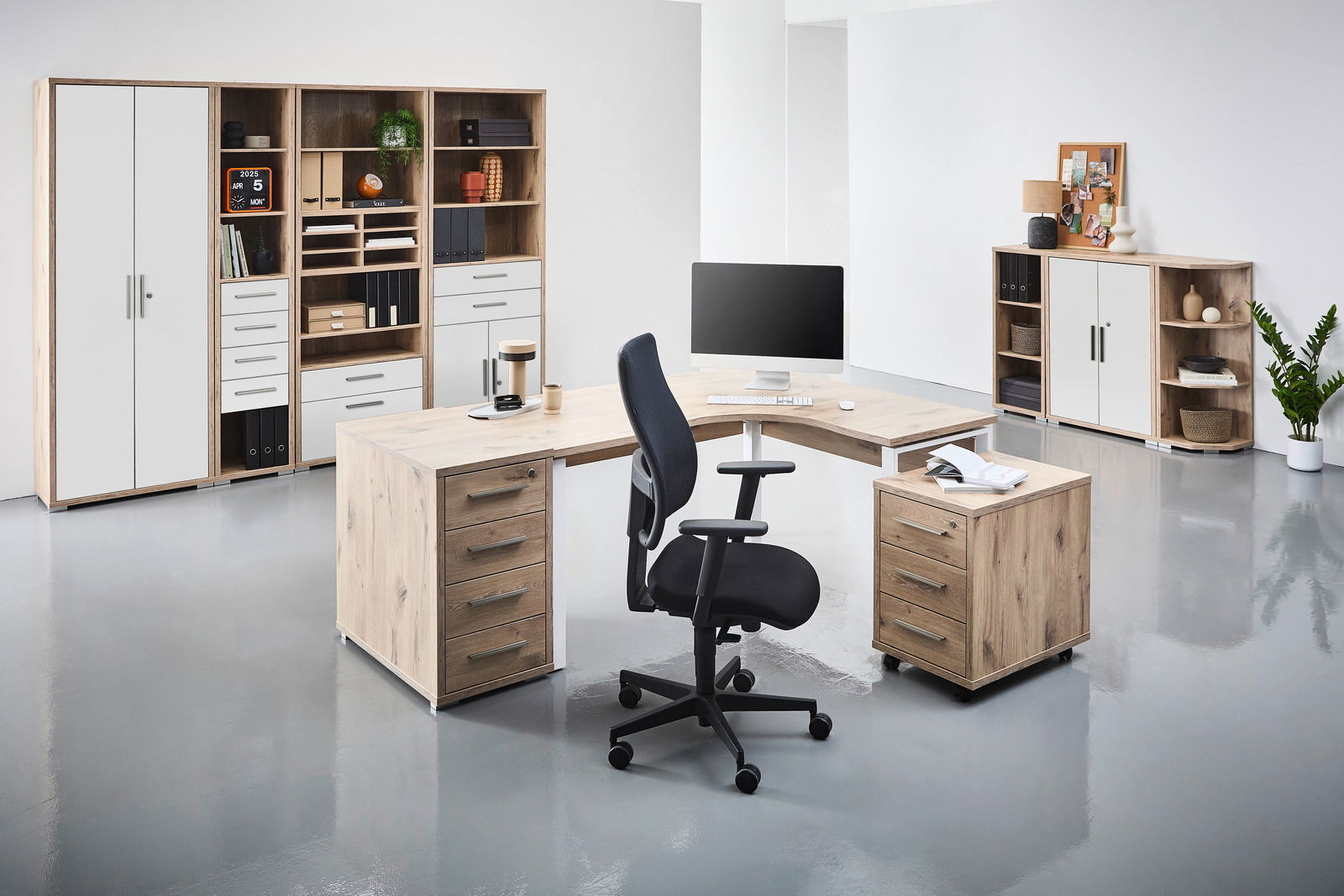 Moderne Büromöbel in einem hellen Raum, bestehend aus einem Eckschreibtisch mit Rollcontainer und einem schwarzen Bürostuhl. Im Hintergrund befinden sich Regale und Schränke mit Aktenordnern und Dekorationen. Perspektive: Frontalansicht.