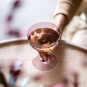 Nahaufnahme einer Hand, die ein Sekt- oder Dessertglas hält, gefüllt mit einem rosafarbenen Getränk, auf einem Holztisch.