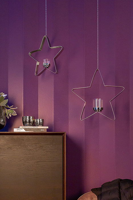 Zwei hängende sternförmige Teelichthalter aus Metall vor einer lila Wand, mit Kerzenhaltern in der Mitte. Perspektive: Frontalansicht.