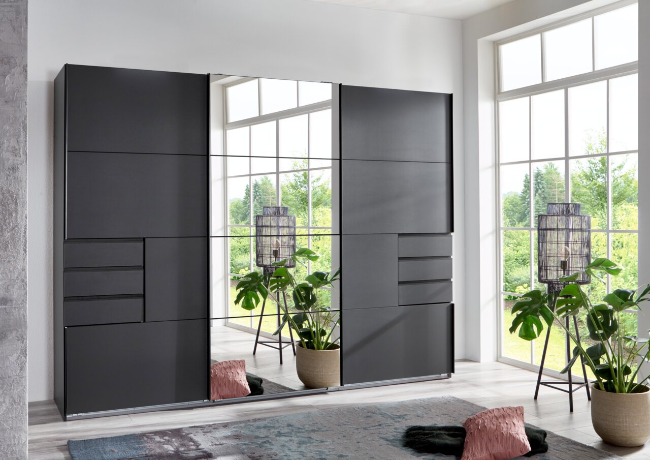 Moderner dreitüriger Schwebetürenschrank in dunklem Design, frontal abgebildet, mit großen Spiegelflächen und Schubladen, umgeben von Pflanzen und dekorativen Elementen in einem hellen Raum.