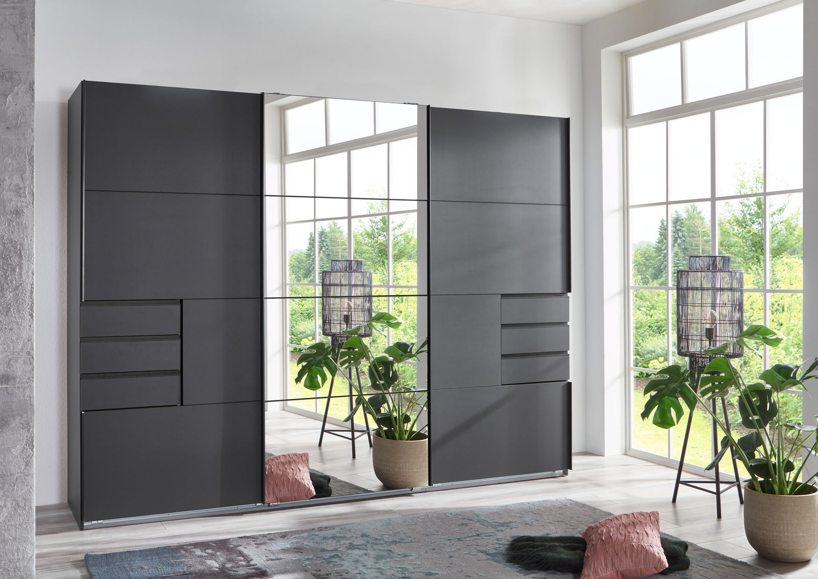 Moderner dreitüriger Schwebetürenschrank in dunklem Design, frontal abgebildet, mit großen Spiegelflächen und Schubladen, umgeben von Pflanzen und dekorativen Elementen in einem hellen Raum.