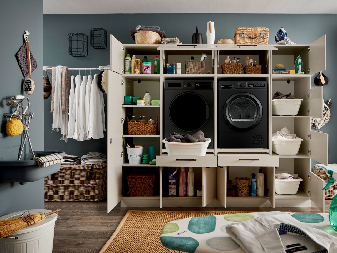 Frontalansicht eines offenen weißen Hauswirtschaftsschranks mit integriertem schwarzen Waschtrockner-Set, Regalböden mit Körben und Reinigungsmitteln sowie Wäschekörben in einem modern eingerichteten Waschraum.