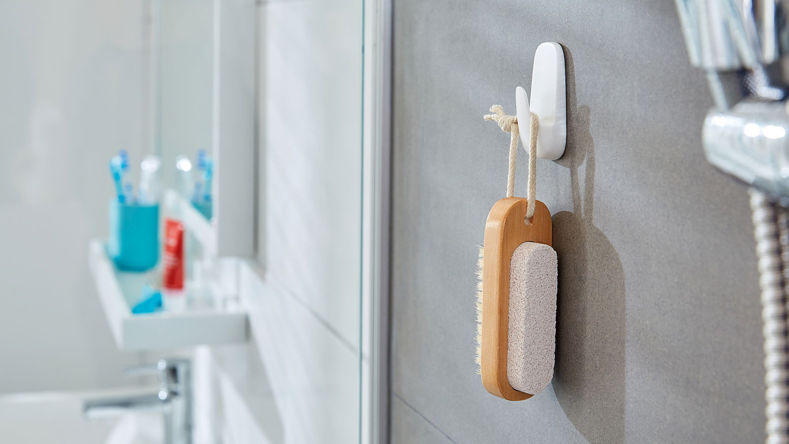 Tesa Powerstrips 2-tlg. WATERPROOF Seitliche Ansicht eines weißen Hakens an einer Badezimmerwand, an dem eine Bürste mit Holzgriff und Bimsstein hängt.