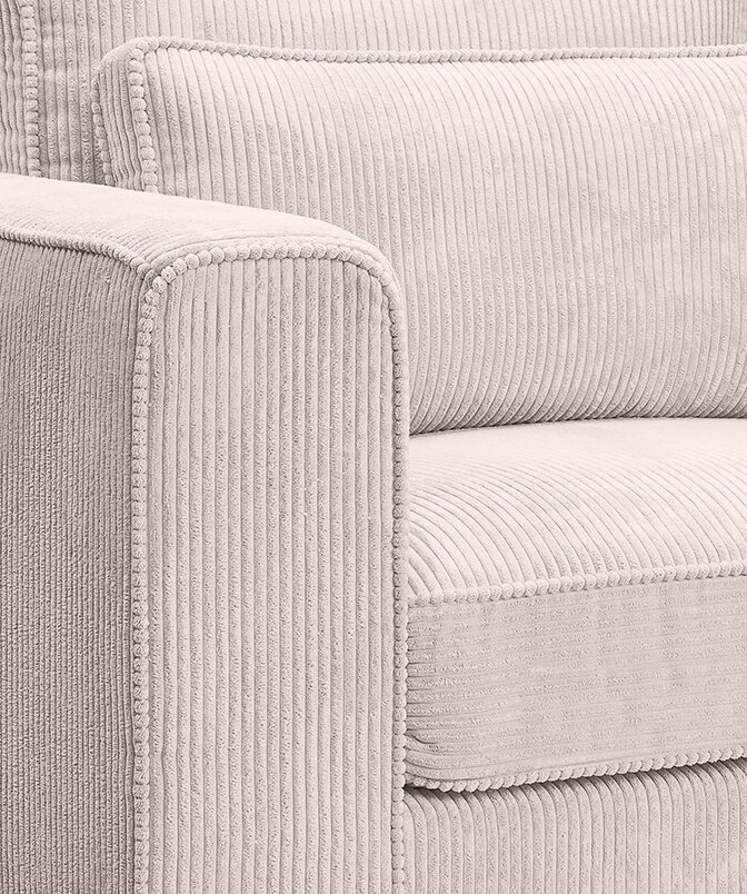 Nahaufnahme eines hellbeigen Cord-Sofas mit breiter Armlehne, Sitz- und Rückenkissen sowie dekorativer Paspelierung, fotografiert aus schräger Vorderseitenperspektive.