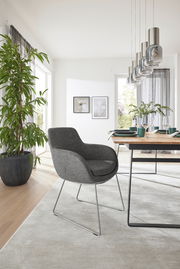 Grauer Armlehnstuhl mit modernem Design, seitliche Perspektive, in einem hellen Esszimmer mit Holztisch und Pflanzen.