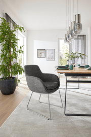 Grauer Armlehnstuhl mit modernem Design, seitliche Perspektive, in einem hellen Esszimmer mit Holztisch und Pflanzen.