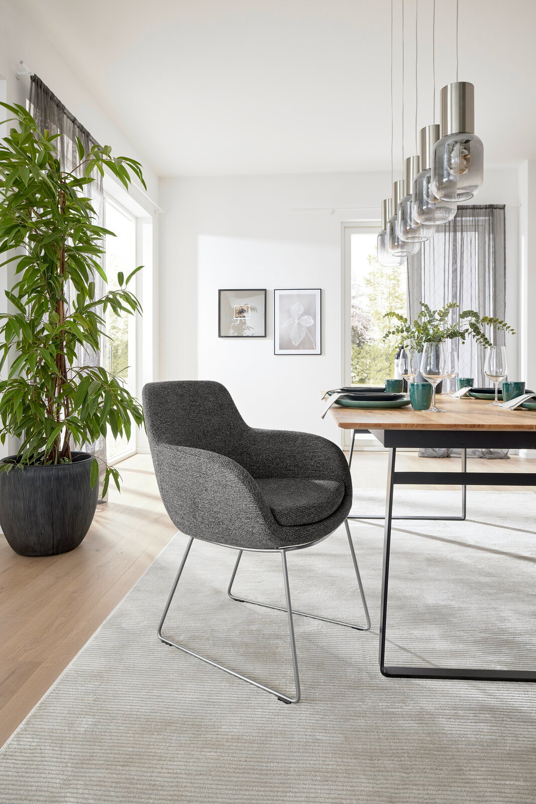 Grauer Armlehnstuhl mit modernem Design, seitliche Perspektive, in einem hellen Esszimmer mit Holztisch und Pflanzen.