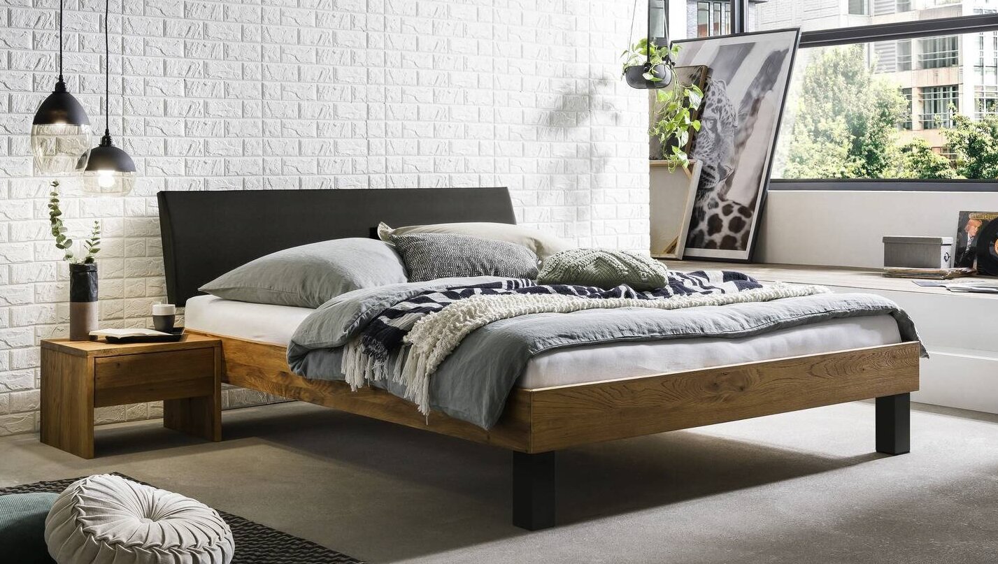 Modernes Holzbett mit schwarzem Kopfteil und grauer Bettwäsche in einem hellen Schlafzimmer mit weißer Ziegelwand und großen Fenstern, Perspektive: leicht schräg von vorne/seitlich.