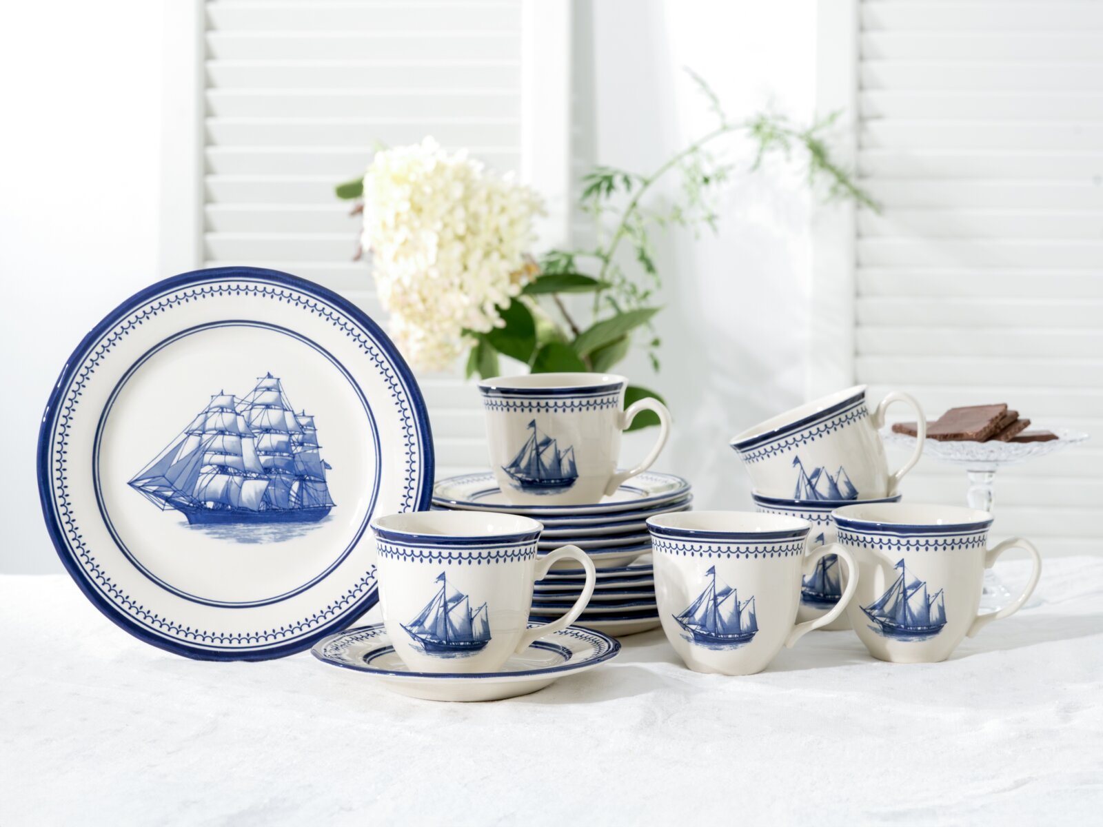 Creatable Kaffeeservice 18-tlg. SEGELSCHIFF Blau-weißes Porzellan-Kaffeeservice mit Segelschiff-Motiv, Tassen, Untertassen und Teller auf weißem Tisch vor hellem Fenster, Frontansicht leicht schräg von vorn.