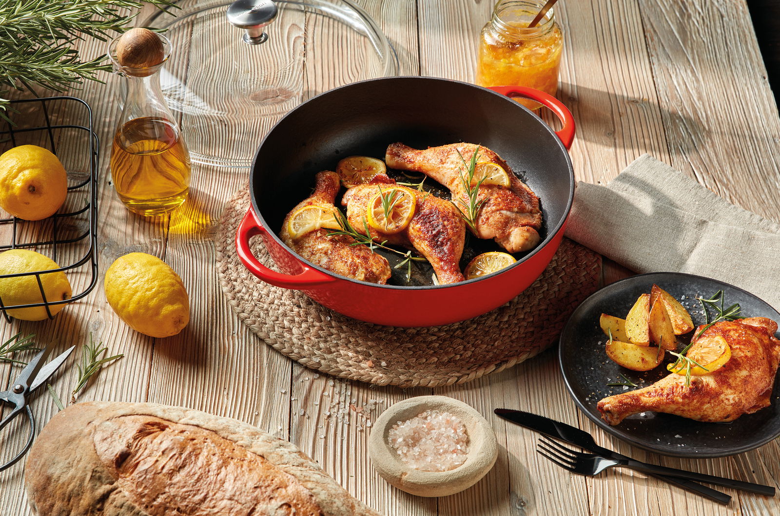 Rote Bauernpfanne Provence 28cm mit gebratenem Hähnchen und Zitronenscheiben, von oben fotografiert, umgeben von frischen Zutaten wie Zitronen, Olivenöl und Brot auf einem Holztisch.