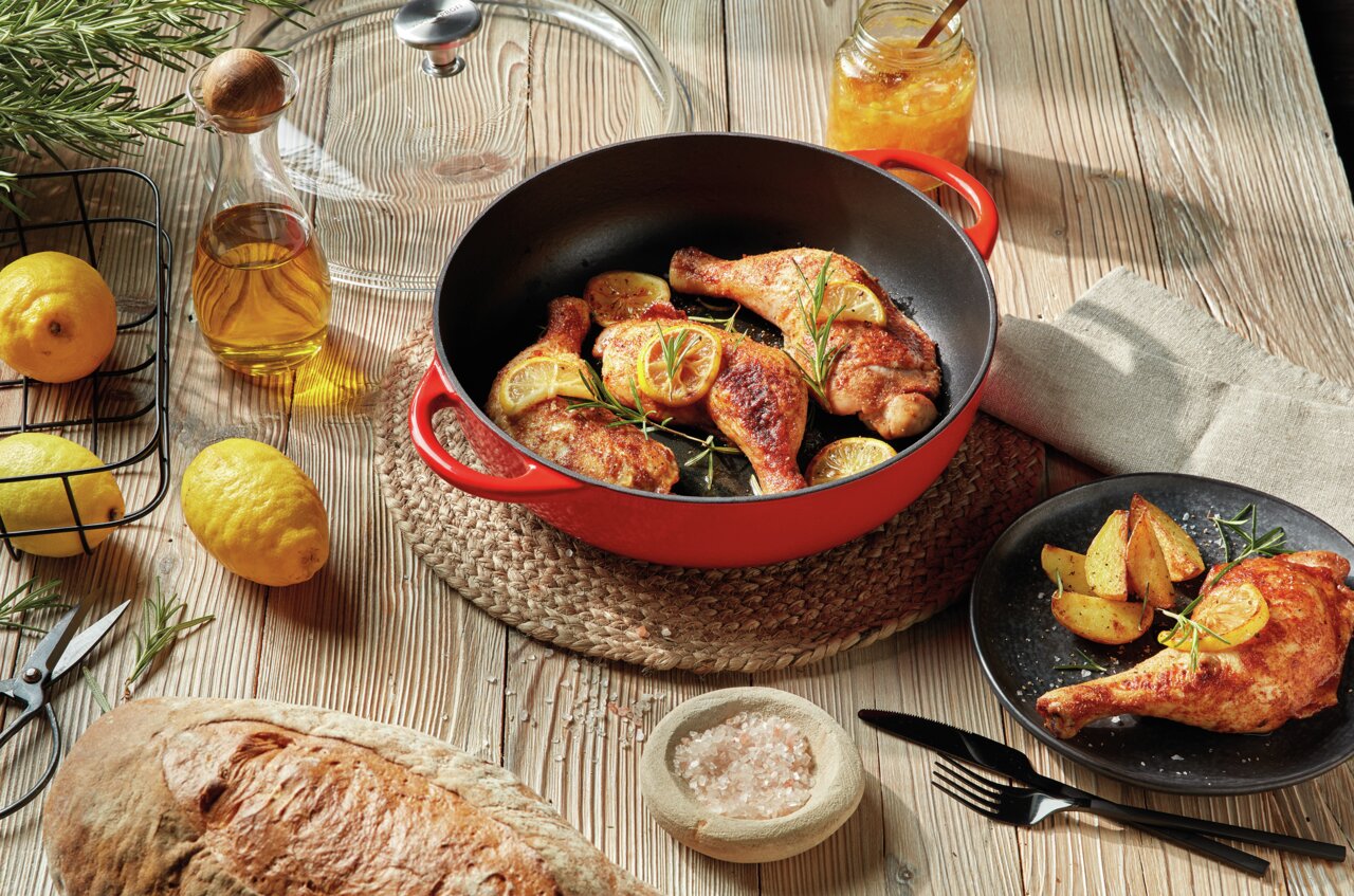 Rote Bauernpfanne Provence 28cm mit gebratenem Hähnchen und Zitronenscheiben, von oben fotografiert, umgeben von frischen Zutaten wie Zitronen, Olivenöl und Brot auf einem Holztisch.