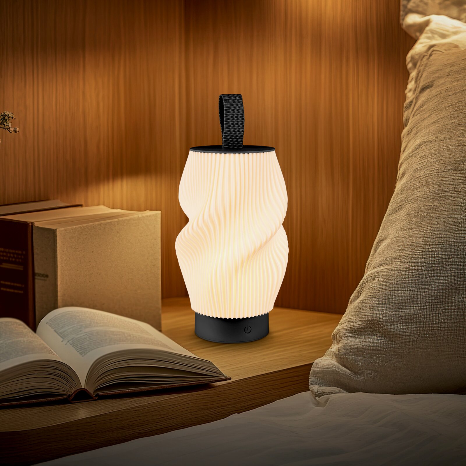 Leuchtende, tragbare LED-Tischlampe mit wellenförmigem, weißem Lampenschirm, schwarzem Sockel und Trageschlaufe auf einem Holz-Nachttisch neben einem offenen Buch, Frontansicht auf Augenhöhe.