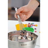 Nahaufnahme eines Rührblitzes PROFI PLUS in einer Hand, der in einer Edelstahlschüssel mit Teig verwendet wird, umgeben von bunten Backformen und Dekorationen.