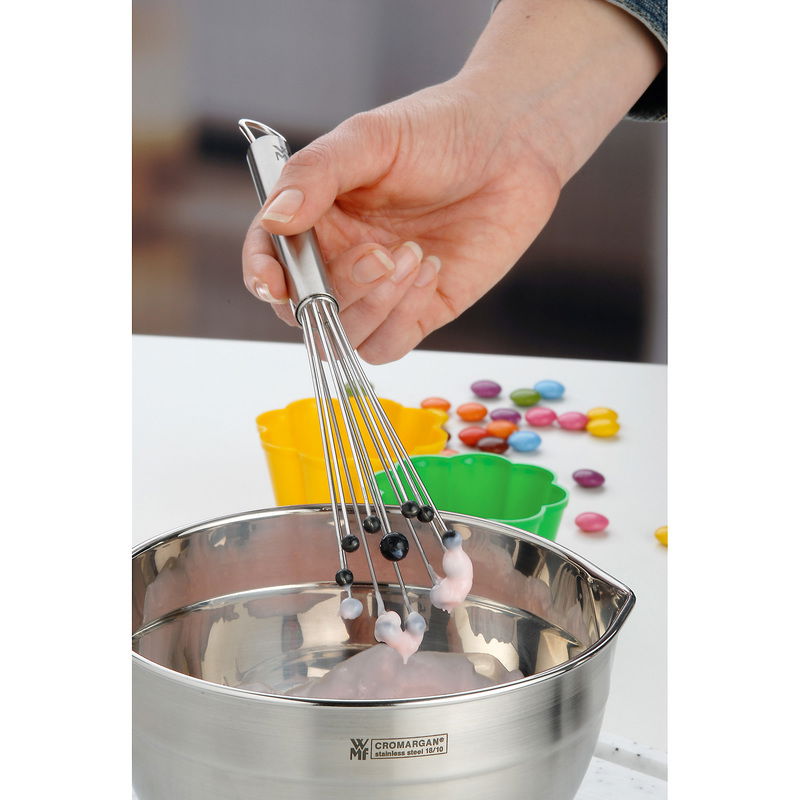 Nahaufnahme eines Rührblitzes PROFI PLUS in einer Hand, der in einer Edelstahlschüssel mit Teig verwendet wird, umgeben von bunten Backformen und Dekorationen.